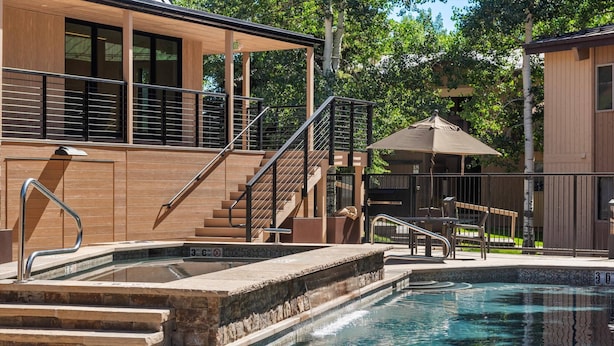 A swimming pool with steps leading into it, a covered seating area, and a building with a wooden facade and a staircase. - Woodbridge 07A (Snowmass Village)