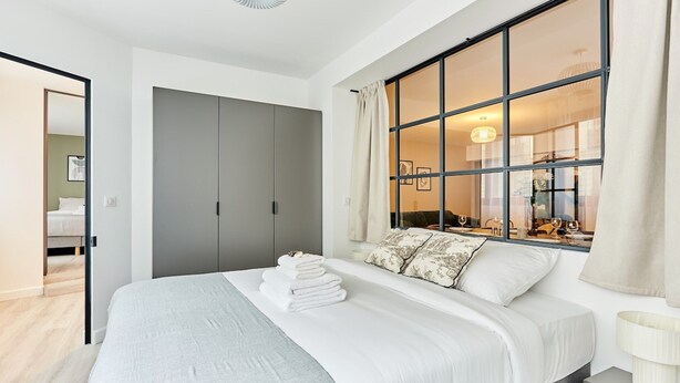 Ein modernes Schlafzimmer mit einem großen Bett, einem grauen Kleiderschrank und einem Fenster mit Blick auf einen Essbereich.