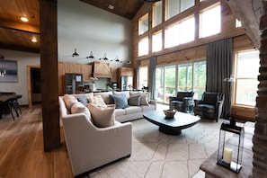 A spacious living room with a large sofa, a coffee table, and a fireplace. There is a kitchen area with wooden cabinets and a refrigerator. Large windows provide natural light and a view of the outdoors.
