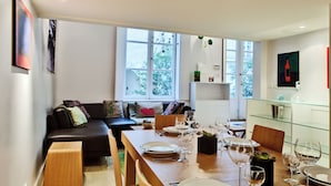 Un soggiorno moderno con zona pranzo, che include un divano, un tavolo in vetro per quattro persone e un'ampia finestra con vista su piante.