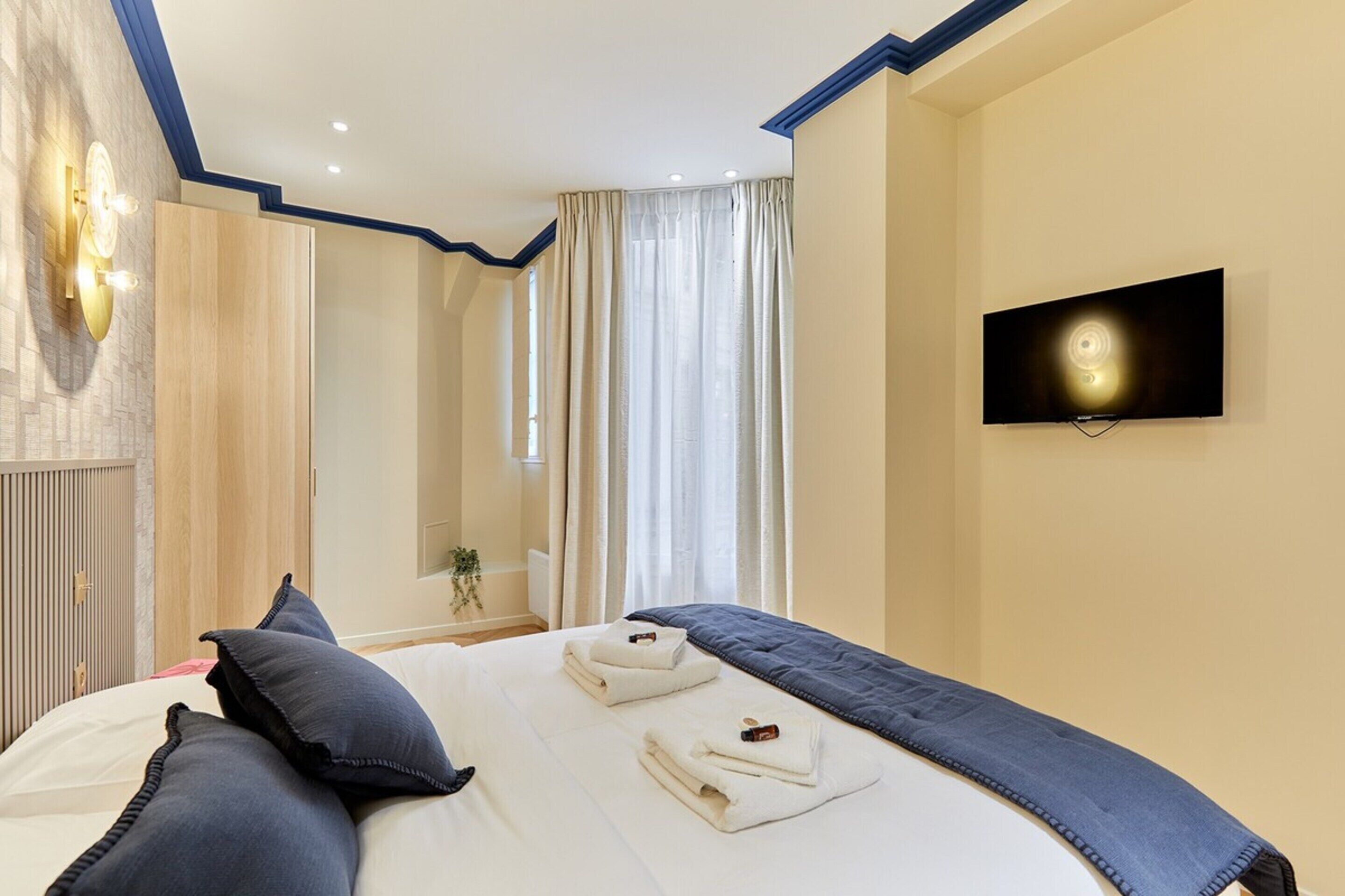 Ein Hotelzimmer mit einem Bett, einem an der Wand montierten Fernseher und einem Fenster mit Vorhängen.
