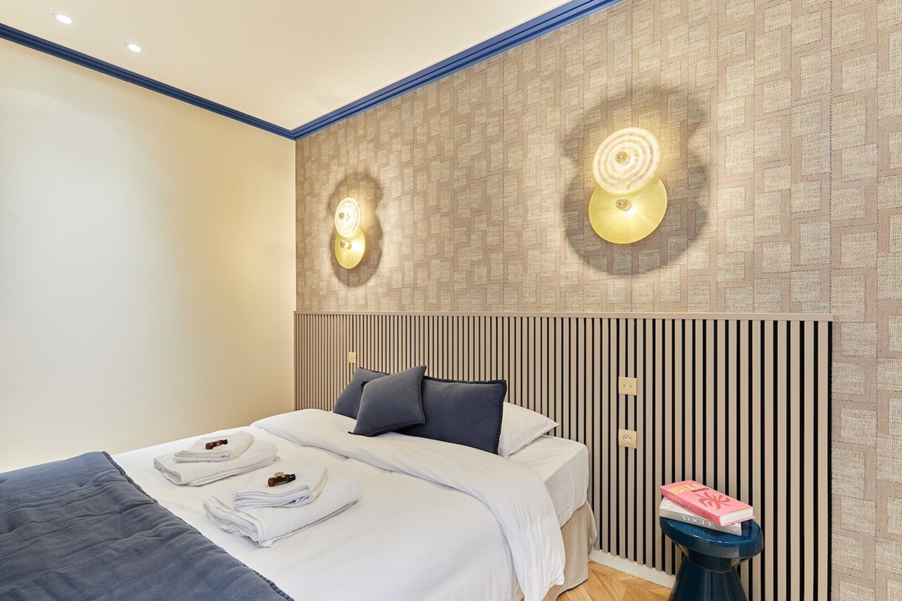 Una habitación de hotel moderna con una cama bien hecha, una mesita de noche con un libro, y una pared con paneles verticales y apliques.