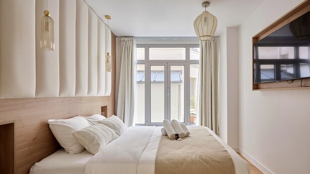 A neatly made bed with white linens and a beige throw blanket, a wooden headboard, and a large window with curtains. - Sublime Flat in the Centre of Paris-montorgueil (Paris)