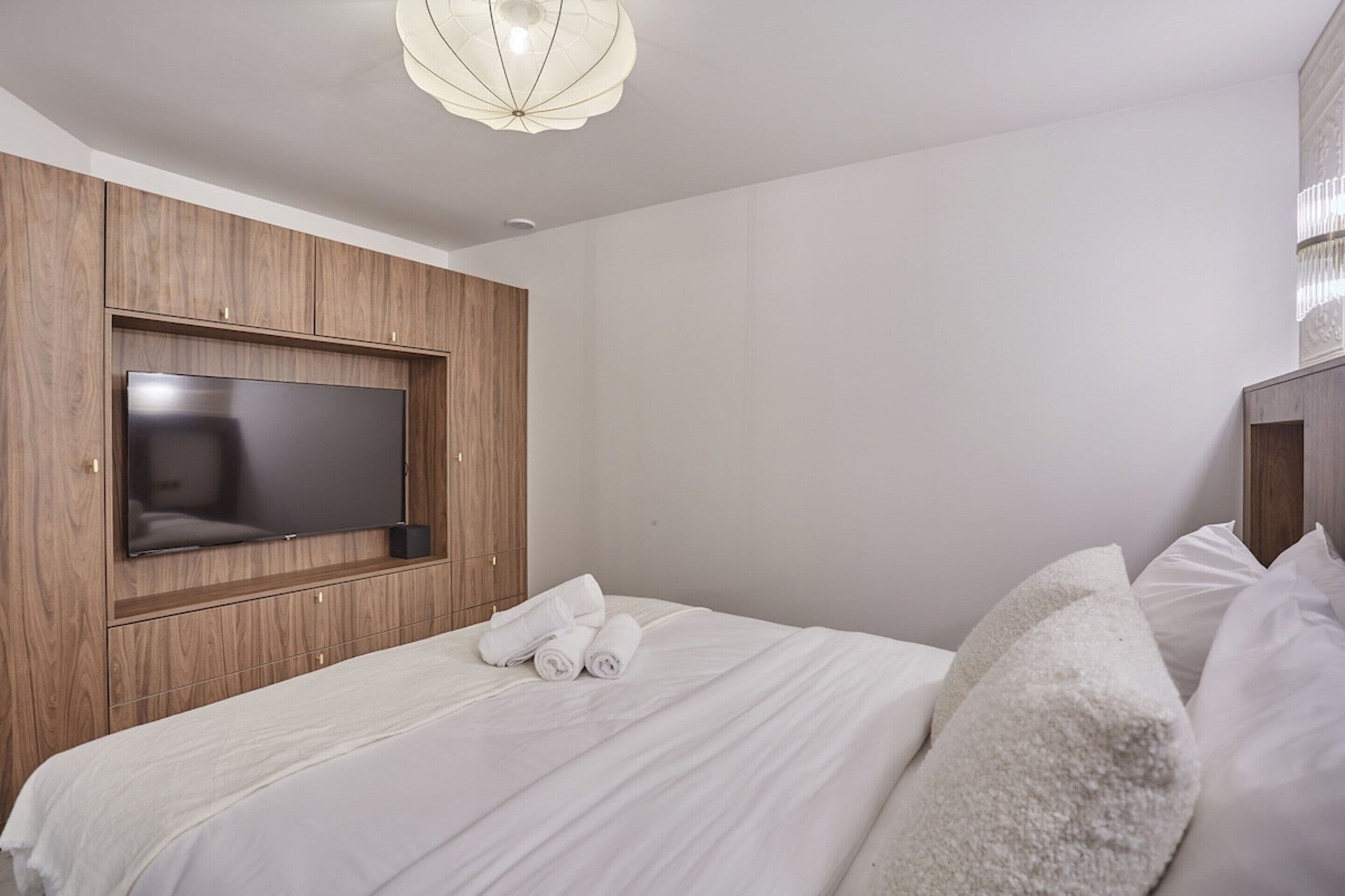 Quarto de hotel moderno com uma cama grande, uma TV de tela plana fixada em um painel de madeira e um lustre no teto.