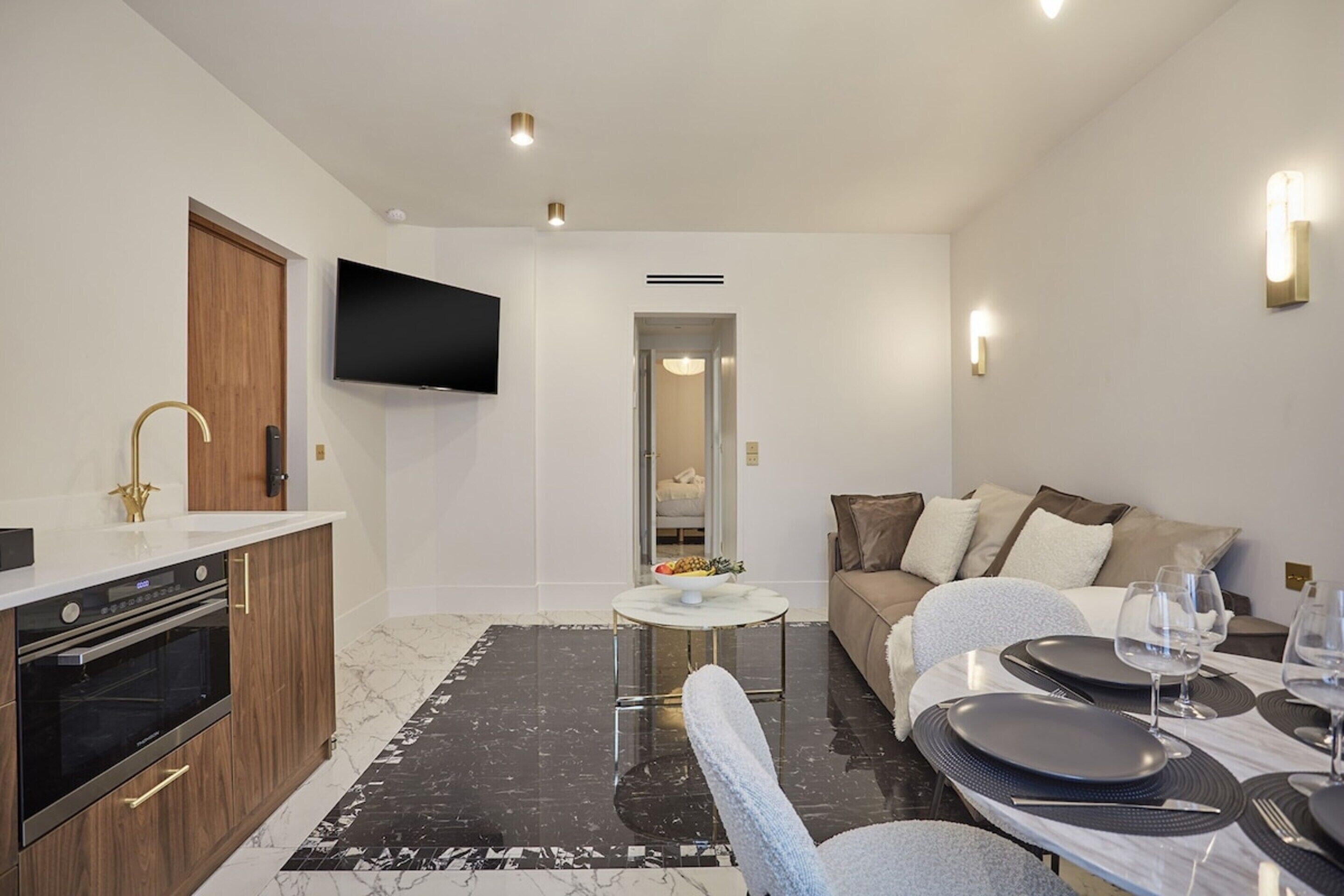 A modern living room with a sofa, a dining table set for two, and a kitchen area with a TV mounted on the wall.