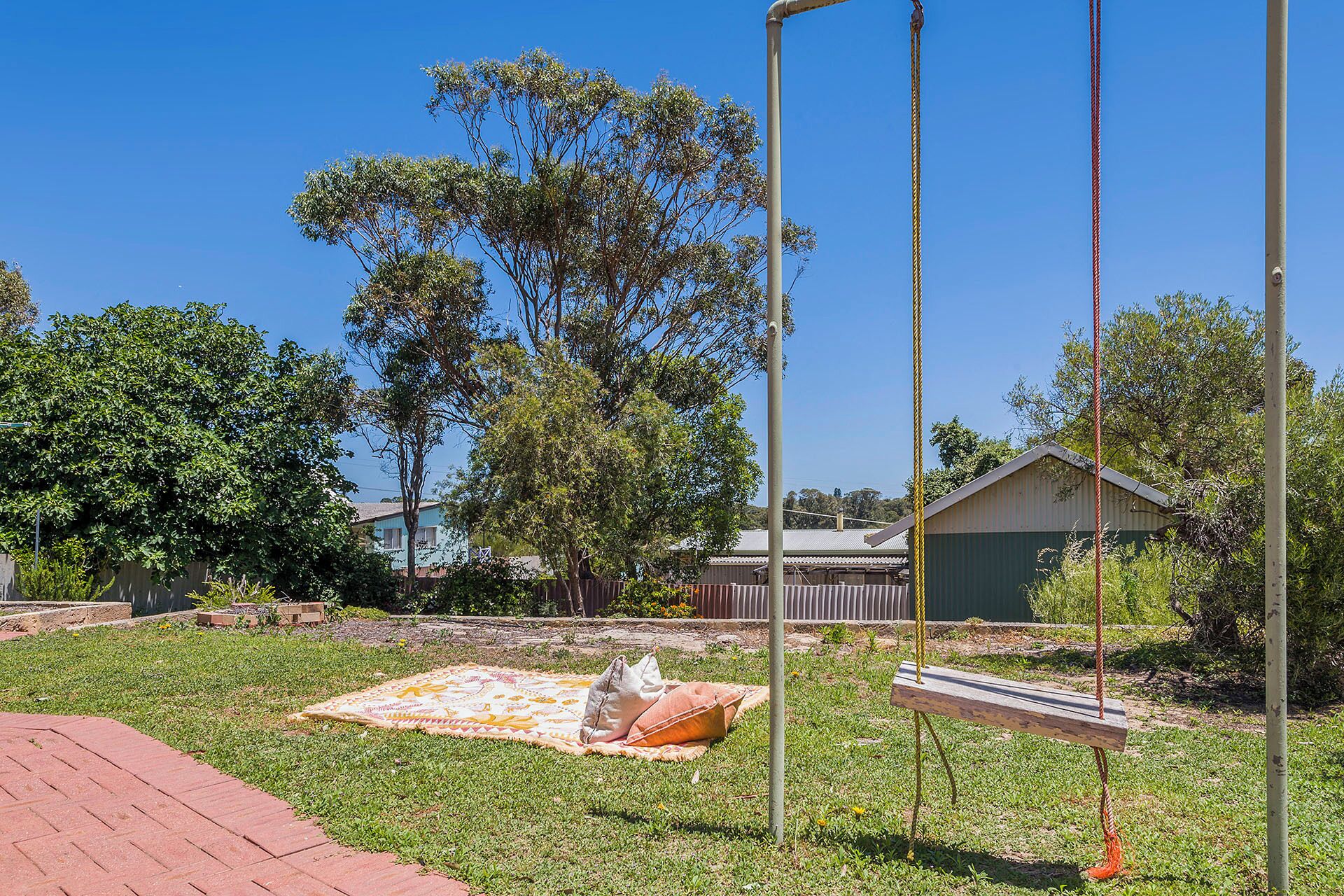 A swing set in a grassy yard with a blanket and pillows on the ground, a wooden swing, and a metal pole.