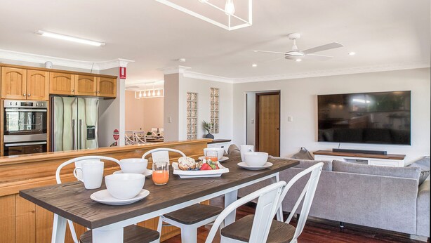 A modern kitchen with built-in appliances, a dining area with a wooden table and white chairs, and a large flat-screen TV mounted on the wall. - Moore than a Shack - Ultimate coastal retreat (Guilderton)