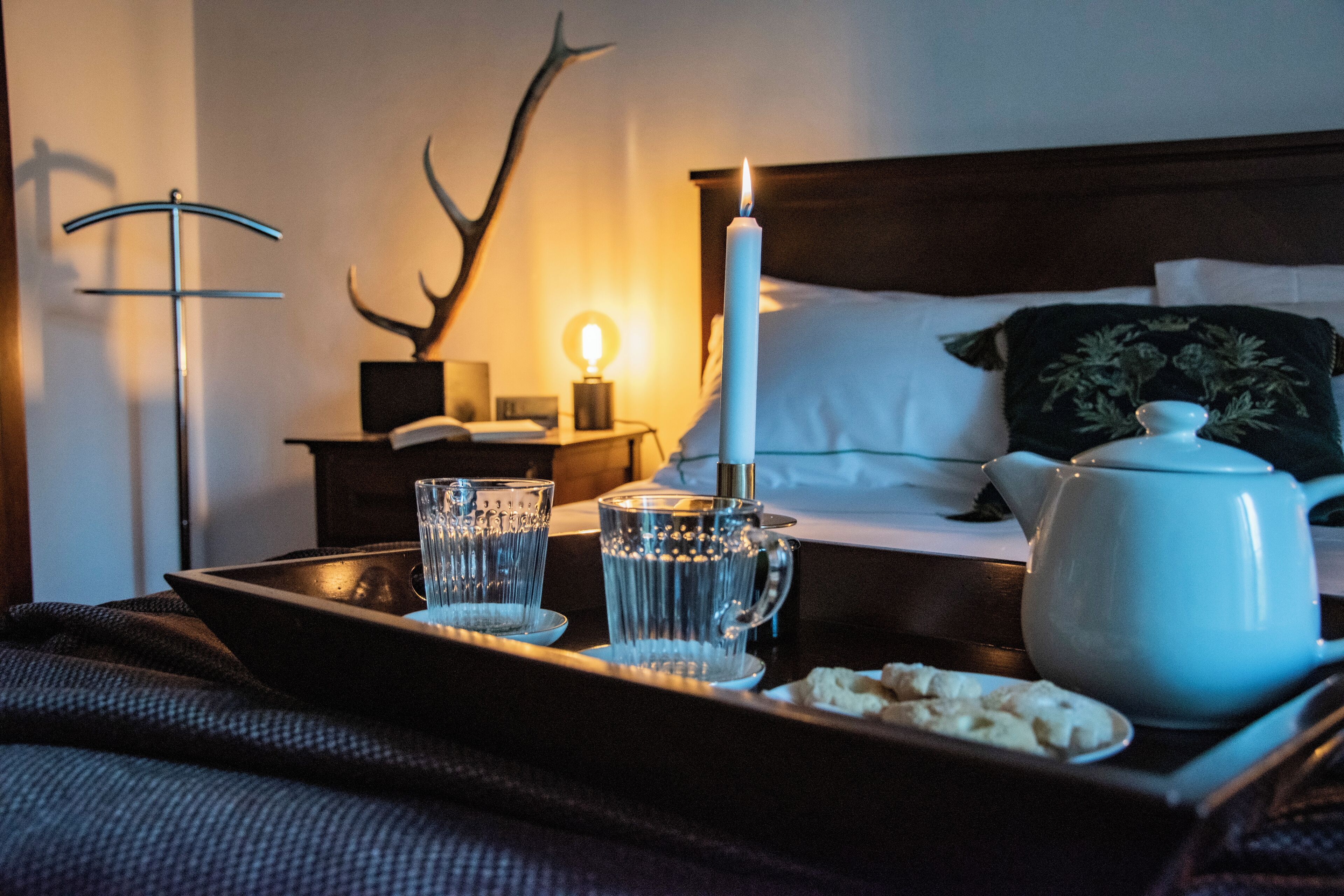 A neatly arranged tray with a white candle, two glasses, and a teapot on a bed.