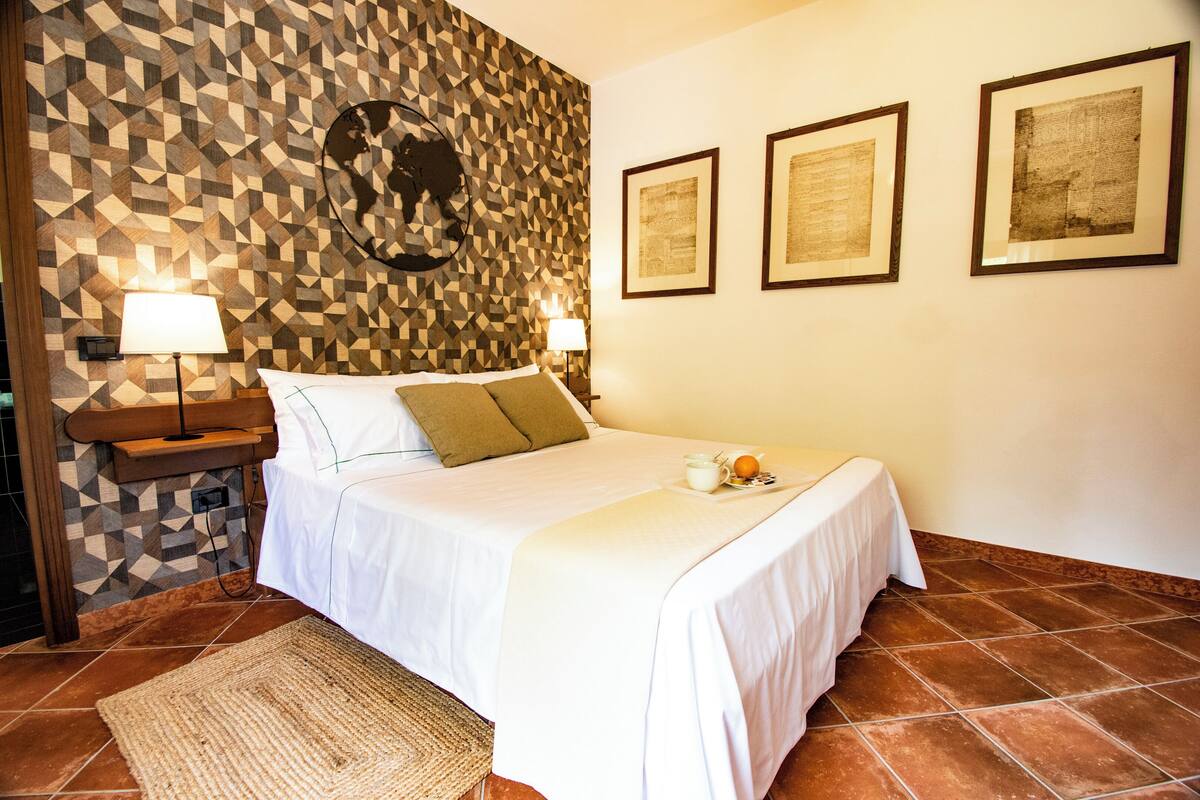 Un lit bien fait, recouvert d’une couvre-lit blanche, avec deux oreillers et un plateau sur lequel se trouvent une tasse et une orange. Deux lampes sont fixées au mur, ainsi qu’une horloge murale et trois tableaux encadrés.