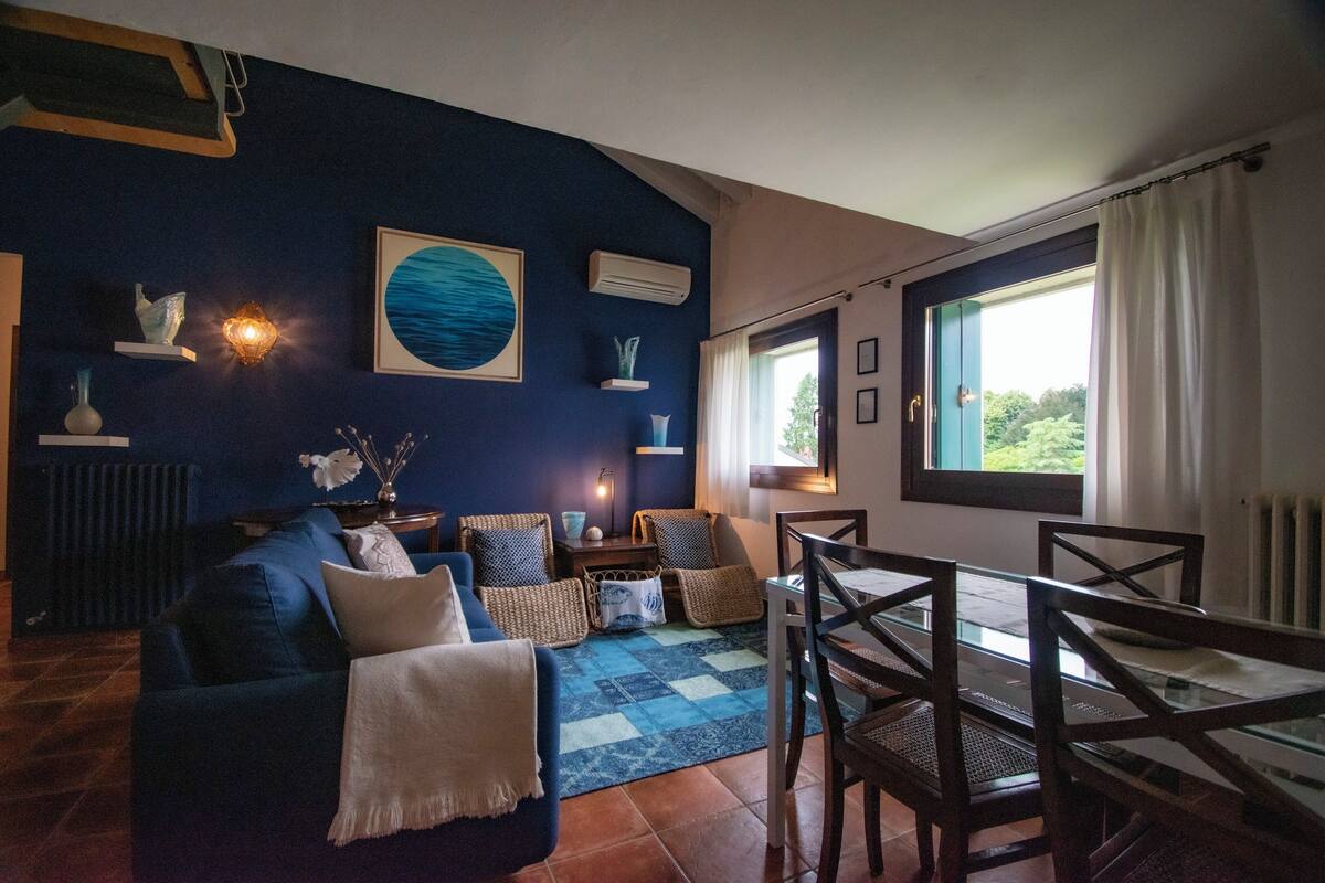 Un salon avec un canapé bleu, une table à manger avec des chaises, un tapis à motifs bleus et blancs, et une vue sur la verdure par une fenêtre.