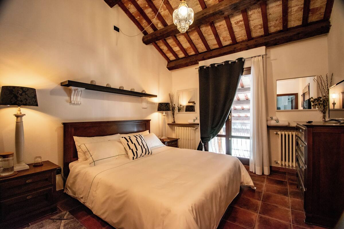 Une chambre à coucher avec un grand lit, une tête de lit en bois, des tables de chevet, une commode, un miroir, un luminaire suspendu et une fenêtre avec des rideaux.