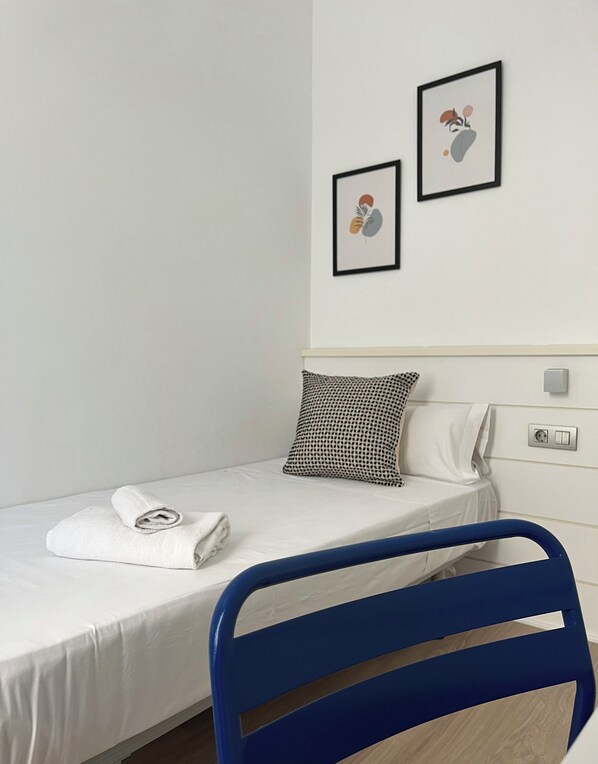 Una cama individual con colchón blanco y una toalla blanca enrollada, una silla de metal azul, un cojín blanco y negro estampado, y dos cuadros en la pared.