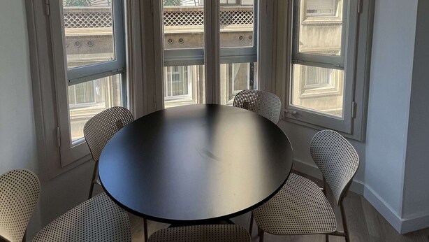 Un comedor con una mesa redonda y seis sillas, ventanas amplias con vistas a edificios, e iluminación empotrada en el techo.