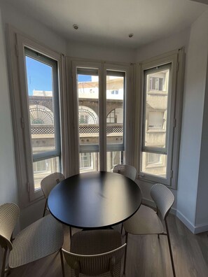 Ein Essbereich mit einem runden Tisch und sechs Stühlen, großen Fenstern mit Blick auf Gebäude und einer Decke mit Einbaulicht.