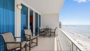 Un balcón con vistas a la playa, que incluye una mesa y sillas, y un ventanal con cortinas azules.