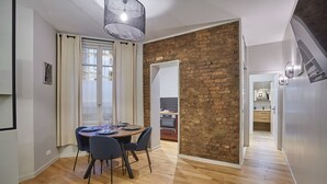 A dining area with a round table, chairs, and a modern pendant light. A brick wall separates the dining area from the kitchen.