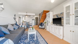 Un salon comprenant un canapé, une table basse et un téléviseur, un escalier menant à la cuisine et un coin repas.