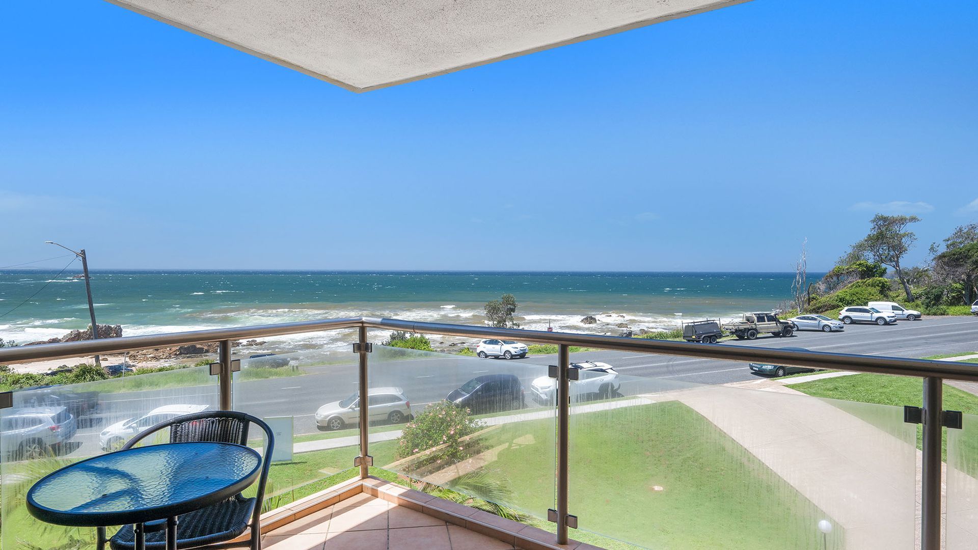 A balcony with a view of the ocean, a road with parked cars, and a grassy area.