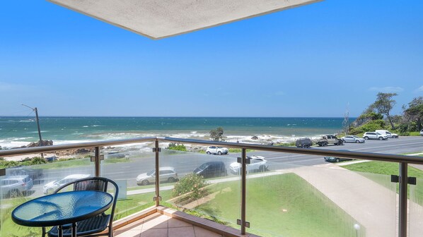 A balcony with a view of the ocean, a road with parked cars, and a grassy area.