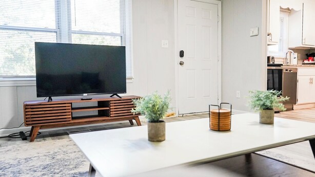 Una sala de estar moderna con un televisor de pantalla plana sobre un soporte de madera, una mesa blanca grande y dos plantas en macetas.
