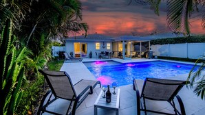 A swimming pool with lounge chairs and a table, surrounded by greenery and a house with an open patio.