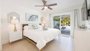 A bedroom with a bed, two bedside tables, a ceiling fan, a large window with a view of the outdoors, and a TV mounted on the wall.