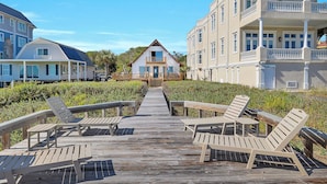 Ein hölzerner Steg führt zu einem Strandhaus mit Balkon, umgeben von üppiger Vegetation und angrenzenden Gebäuden.