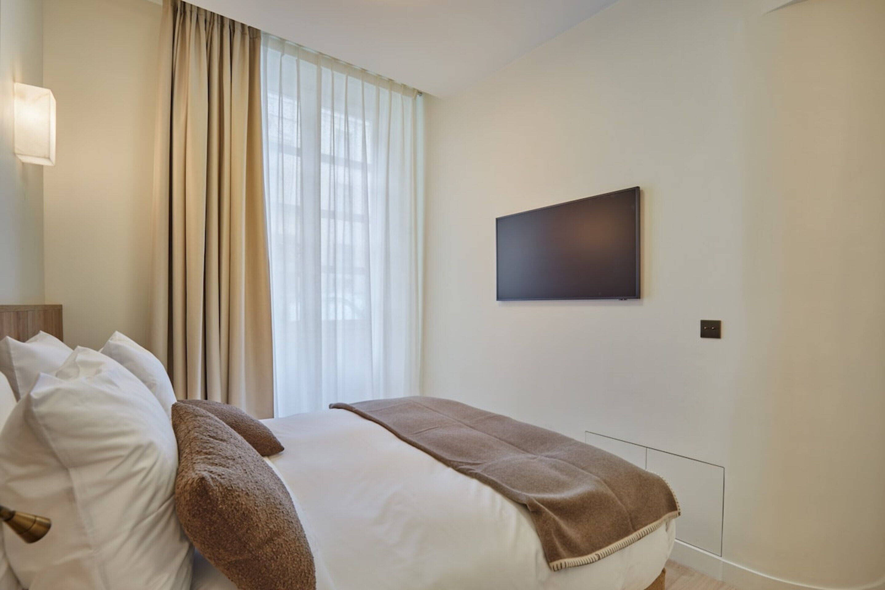 A hotel room with a bed, a flat-screen TV mounted on the wall, and a window with curtains.