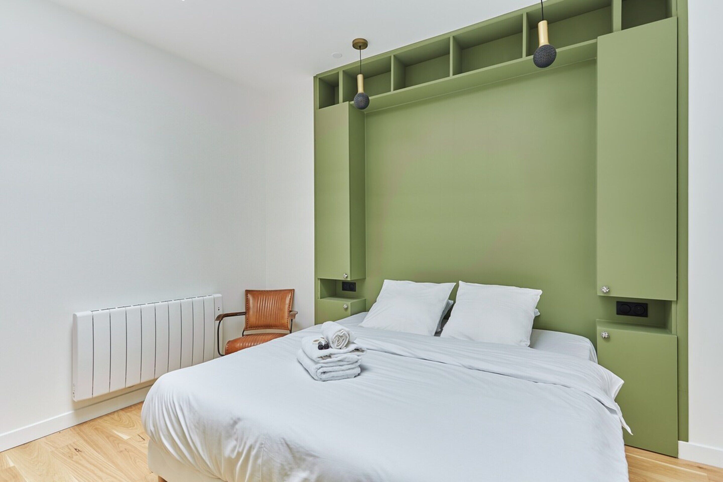 Ein modernes Schlafzimmer mit einem grünen Kopfteil, einem ordentlich bezogenen Bett mit weißen Leinen, einem Holzstuhl und einer Heizung.