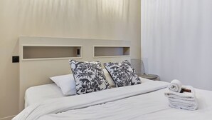 Un lit bien fait, avec du linge de lit blanc et deux coussins à motifs floraux, une serviette pliée posée sur le côté, et une table de chevet avec une lampe.