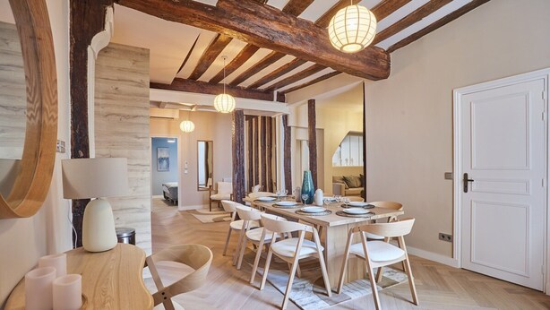 Un comedor con una mesa de madera dispuesta para seis personas, rodeada de seis sillas blancas. Se ve una mesita de madera con velas a la izquierda. La habitación tiene vigas de madera a la vista en el techo y una puerta blanca a la derecha.