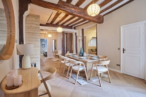Ein Essbereich mit einem Holztisch, gedeckt für sechs Personen, umgeben von sechs weißen Stühlen. Ein Nachttisch aus Holz mit Kerzen ist links sichtbar. Der Raum zeichnet sich durch freiliegende Holzbalken an der Decke und eine weiße Tür rechts aus.