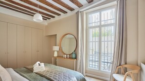 Un dormitorio con cama, mesita de noche, espejo, ventana con cortinas y una silla de madera.