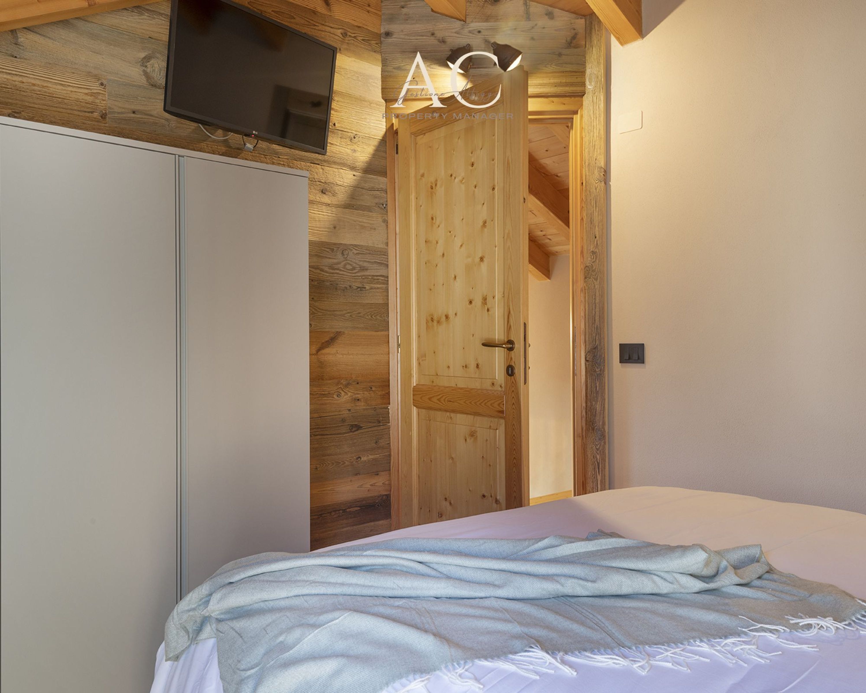 Ein Schlafzimmer mit Holztür, einem Bett mit weiß-blauer Bettdecke und einem an der Wand befestigten Fernseher.
