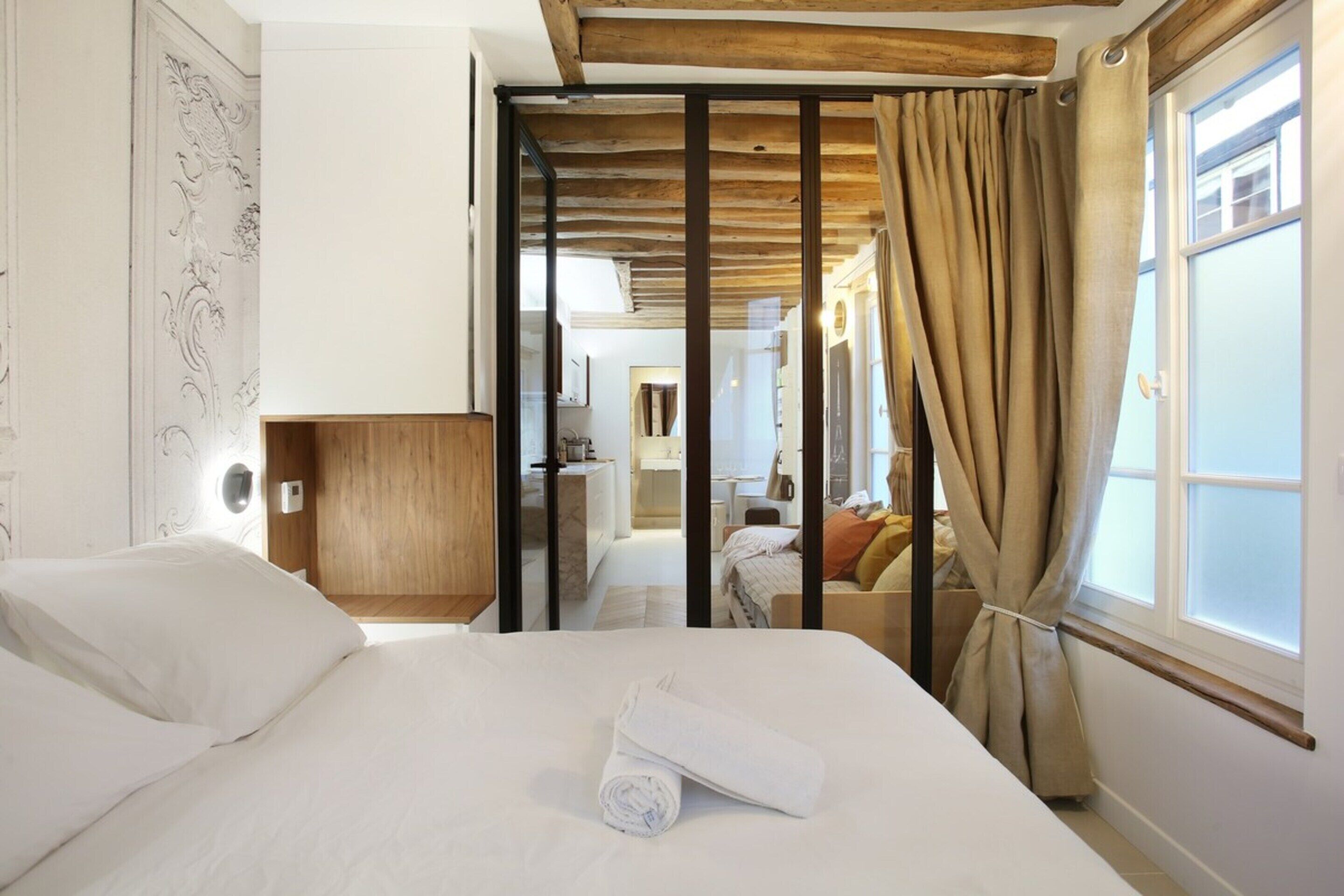 A hotel room with a bed, a wooden headboard, a window with curtains, and a view of the bathroom and kitchen area.