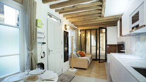 A modern kitchen with white cabinets, a marble countertop, and a wooden ceiling. There is a dining area with a table set for four, and a bed in the background. - Superb Apartment Place des Vosges Le Marais (Paris)