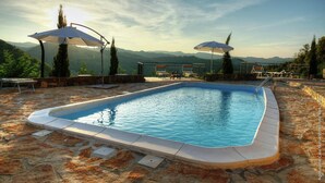 A swimming pool with stone flooring, surrounded by umbrellas and a scenic mountain view.