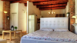 A bedroom with a large bed, a wooden table, a refrigerator, and a brick wall.