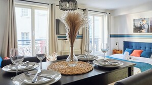 A hotel room with a dining table set for two, a bed with a blue headboard, and a view of the city through large windows. - Cosy Apartment Le Marais Center of Paris (Paris)