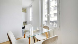 Un comedor con una mesa de vidrio y cuatro sillas, un ventanal con vista a un edificio y una pared decorada al fondo.