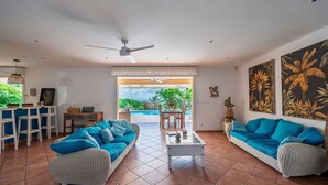 Un vaste espace de vie comprenant deux canapés, une table basse et un coin repas avec des chaises. On peut admirer une piscine et de la verdure depuis l’intérieur.