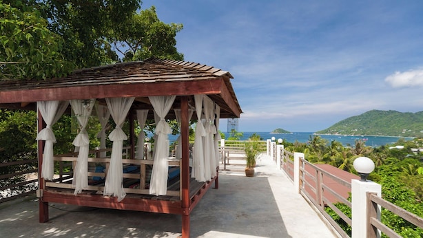 A wooden gazebo with white curtains, a concrete pathway, and a view of the sea and hills.