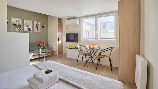 A modern hotel room with a bed, a small dining area with a table and chairs, a sofa, a TV, and artwork on the walls.