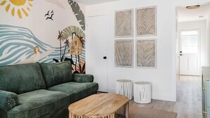 Una sala de estar con un sofá verde, una mesa de centro de madera y un mural que muestra una escena de playa. Hay cuatro obras de arte enmarcadas en la pared y dos taburetes redondos.