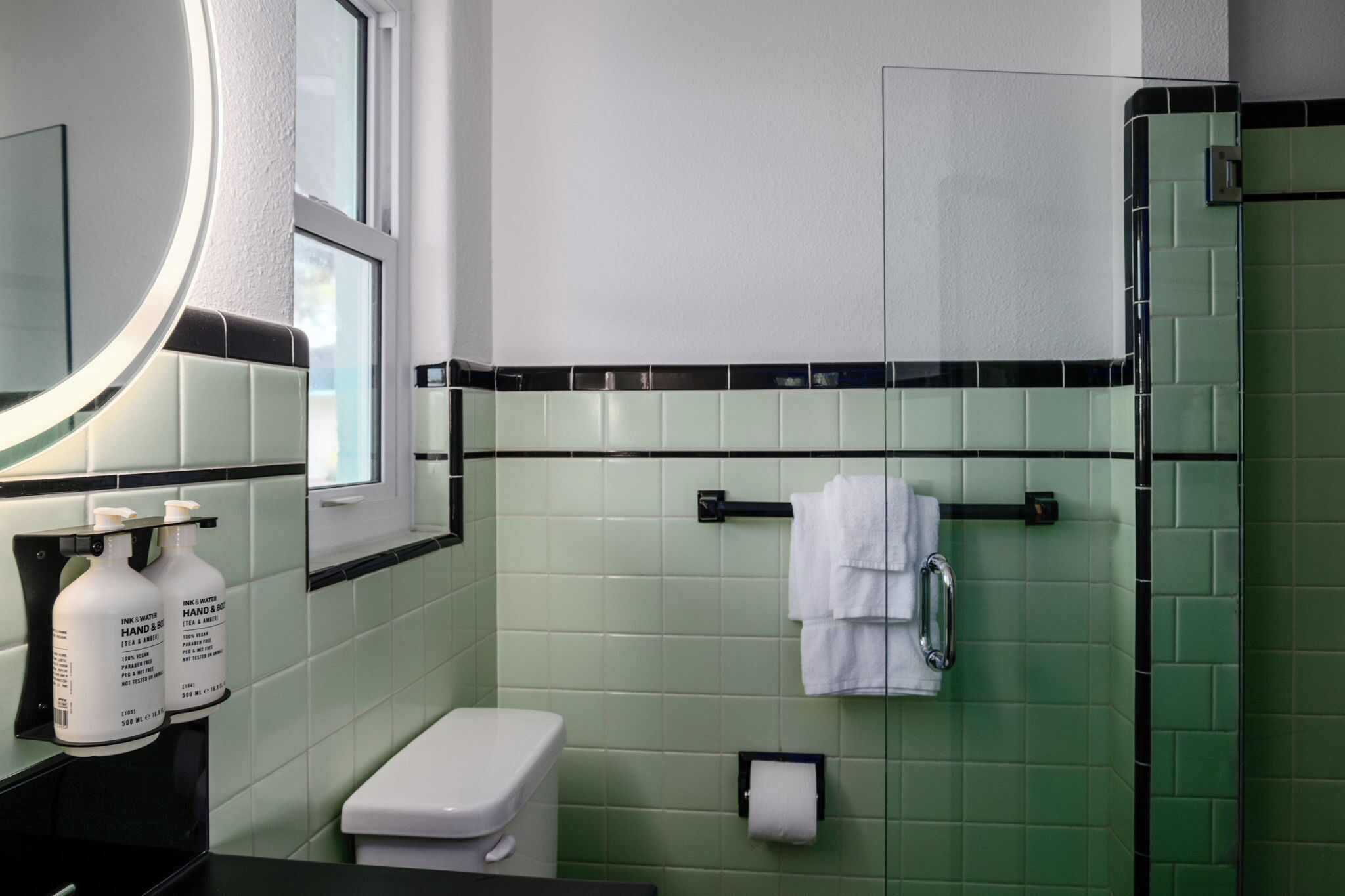 A bathroom with green tiles, a white toilet, a sink with hand soap, and a mirror.