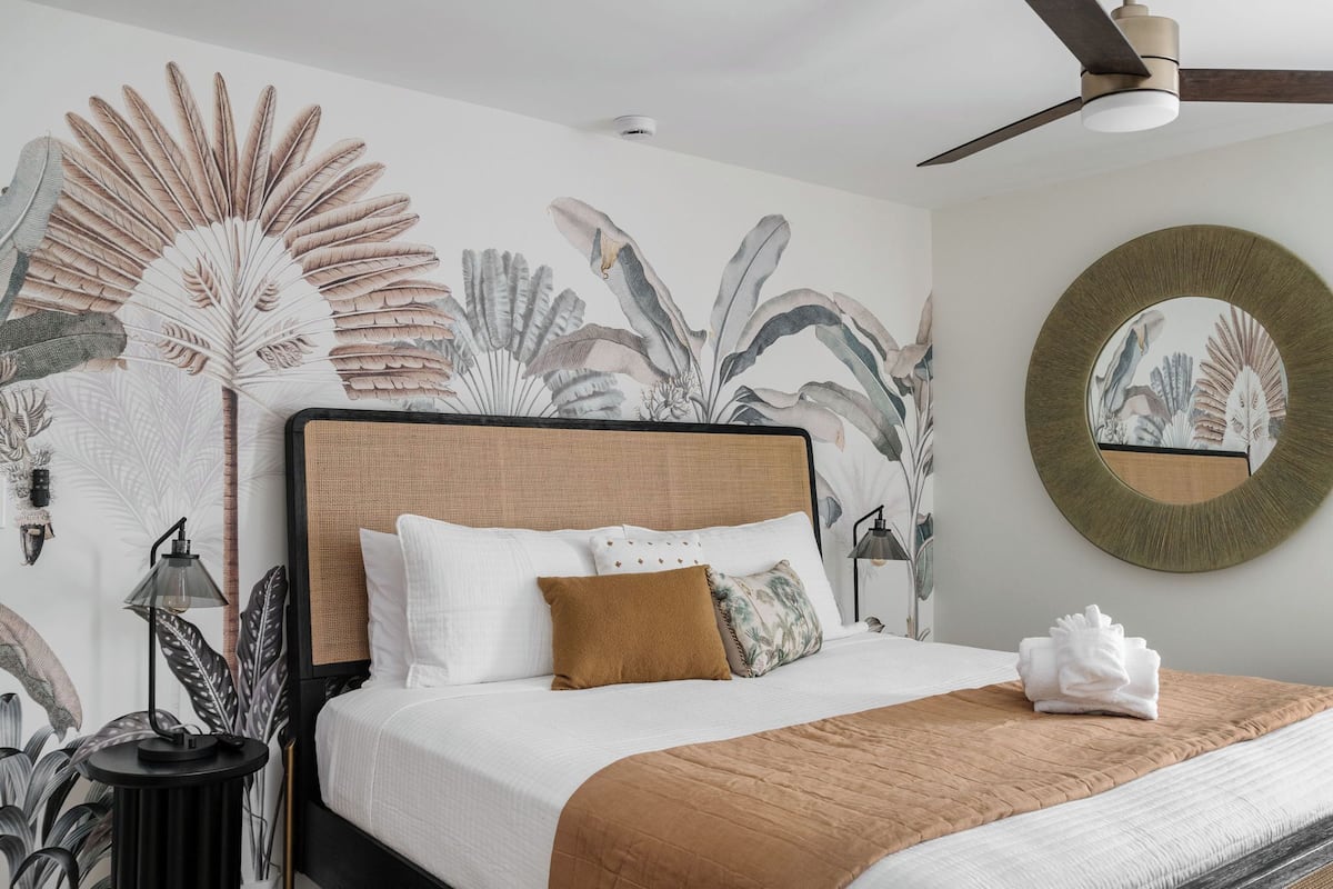 Una cama bien hecha con una manta marrón, almohadas blancas y un plaid marrón. Hay un espejo redondo en la pared y un ventilador de techo sobre la cama.