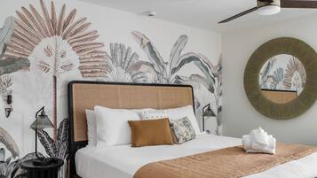 A neatly made bed with a brown blanket, white pillows, and a brown throw. There is a round mirror on the wall and a ceiling fan above the bed.