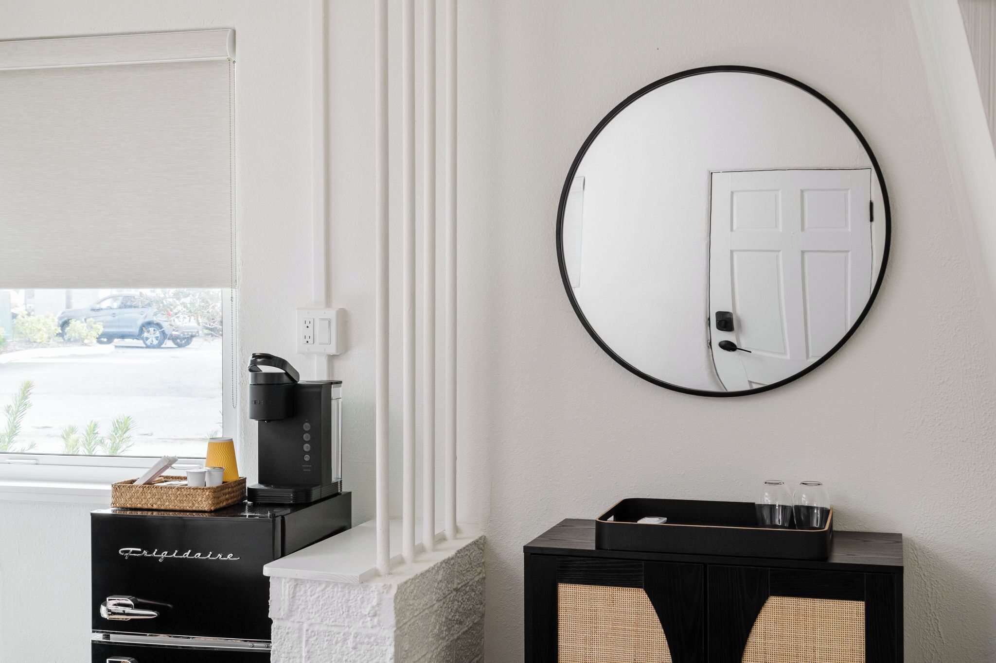 Een moderne hotelkamer met een open haard, een koffieapparaat, een zwarte console tafel met glazen en een ronde spiegel aan de muur.