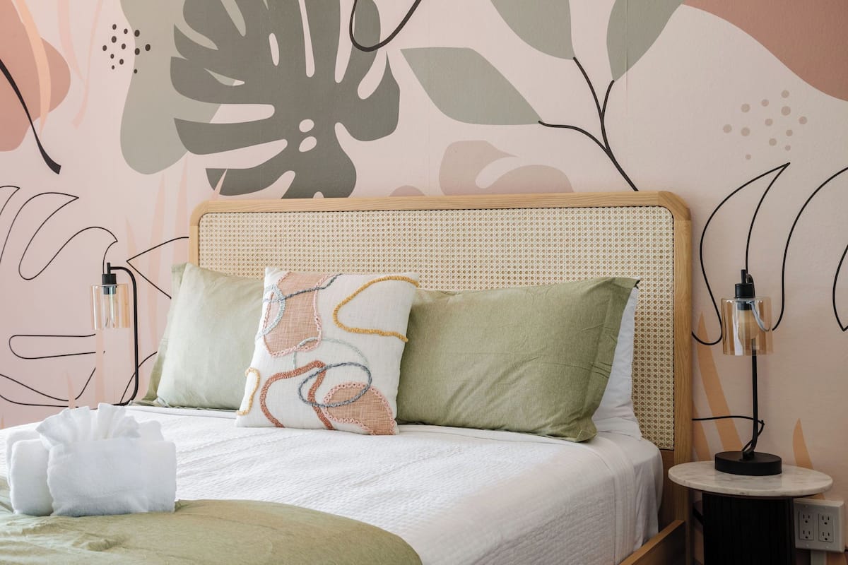 Una cama bien hecha con cabecera de ratán, dos almohadas, una lámpara de mesita de noche y una mesita redonda con lámpara.