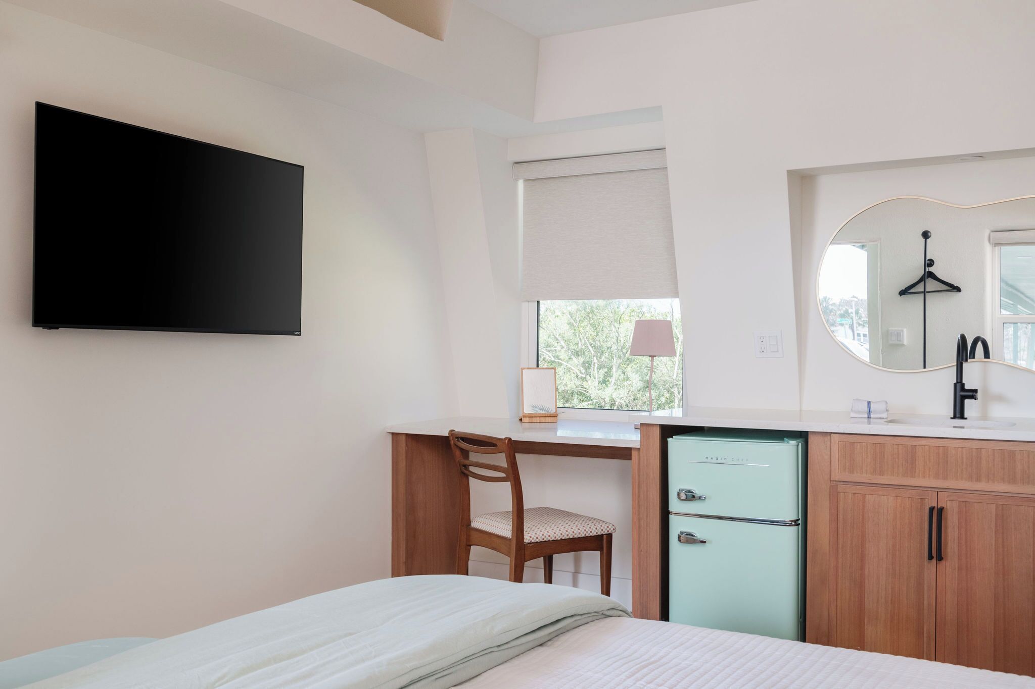 A bedroom with a bed, a desk with a chair, a small refrigerator, a TV mounted on the wall, and a window with a view of trees.